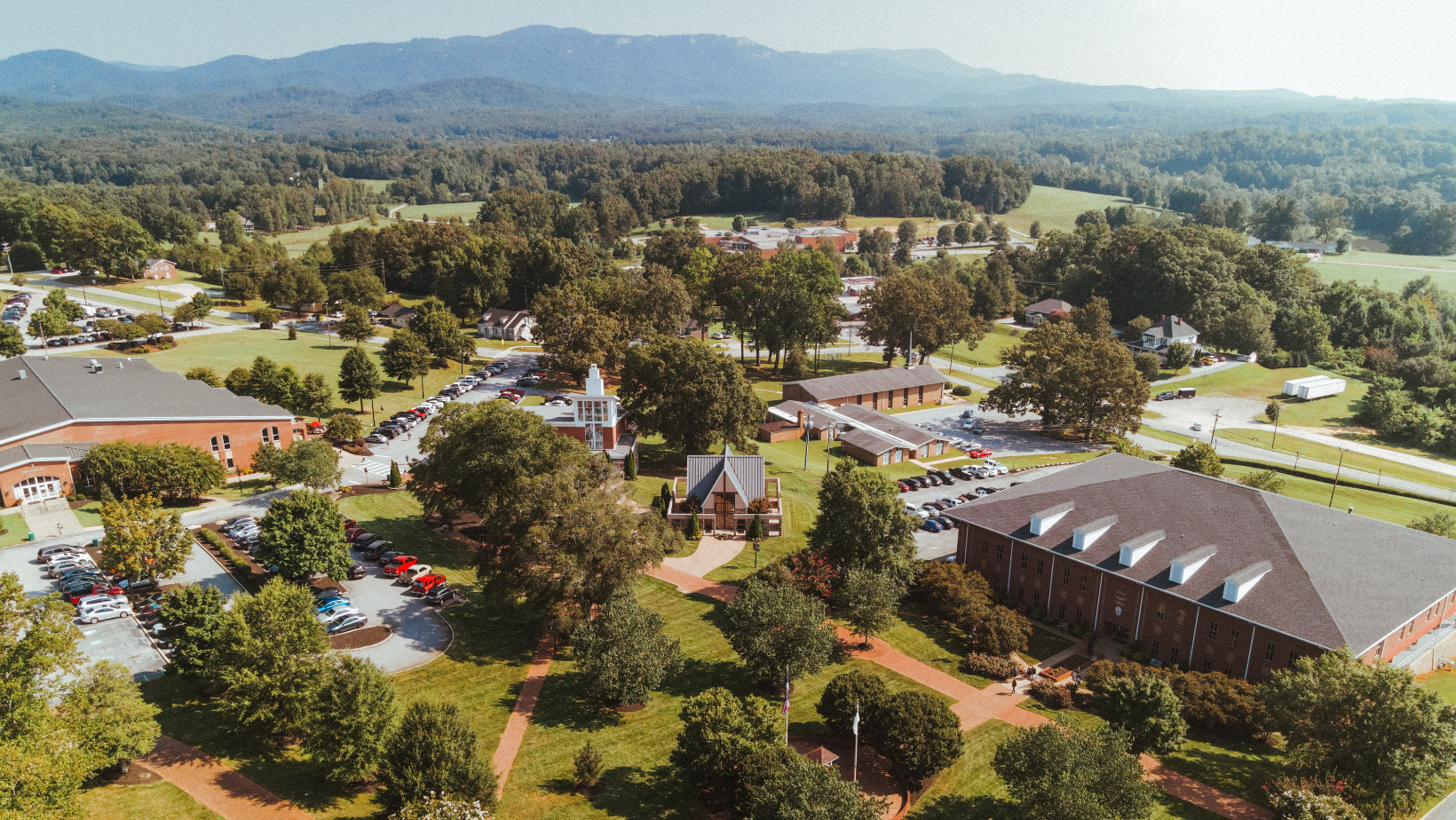 Aerial view of a campus with buildings, parking lots, and trees with mountains in the background.