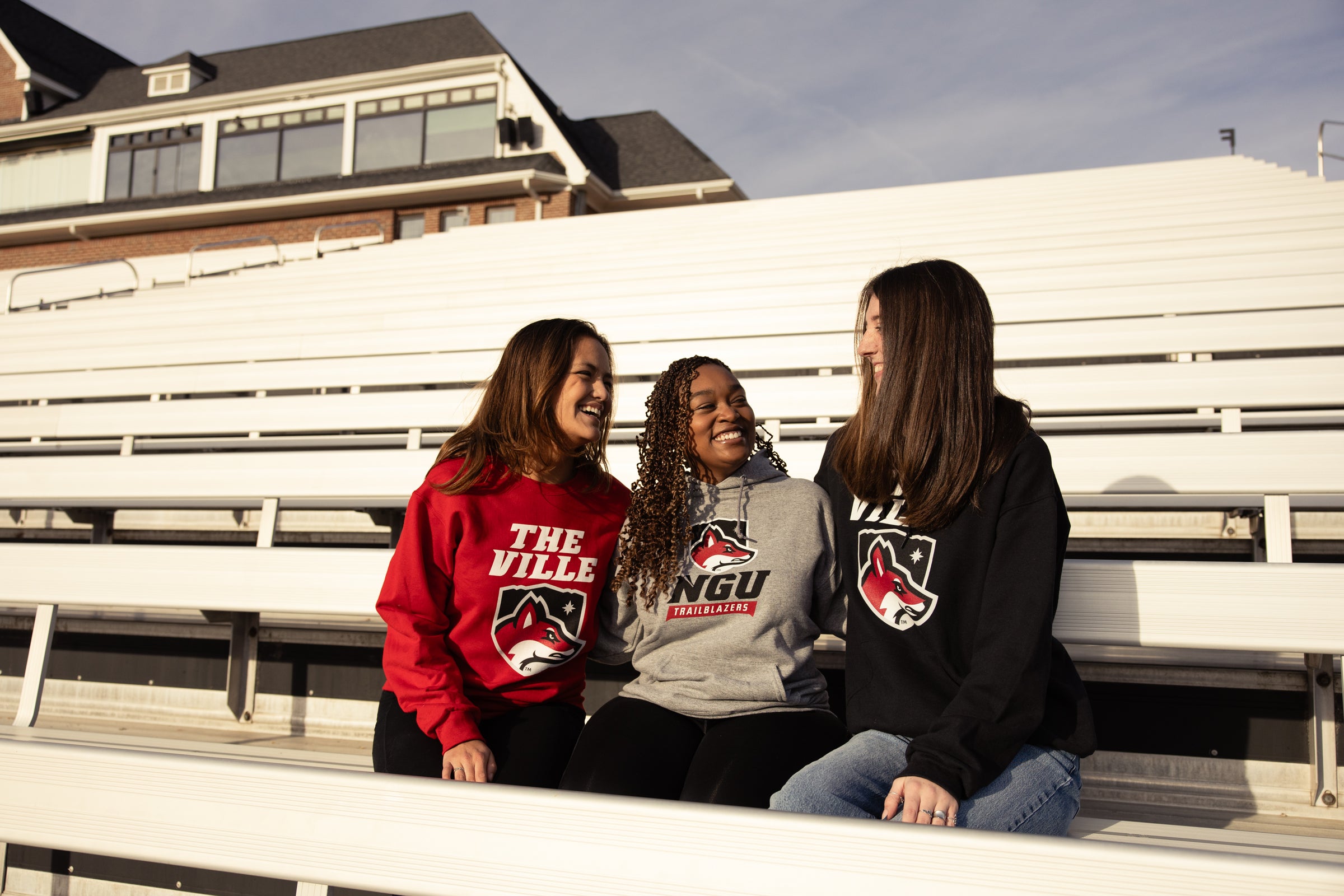 Three women sitting on bleachers wearing sweatshirts with NGU logos.